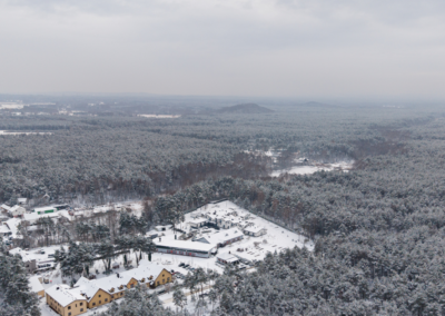 Widok z lotu ptaka na ośnieżony Hotel Jurajski Kryształ zimą, otoczony gęstym, przyprószonym śniegiem lasem sosnowym. W oddali widać zarysy skałek Jury Krakowsko-Częstochowskiej.