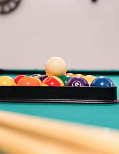 Szczegółowe zdjęcie zestawu kolorowych bil bilardowych ułożonych w czarnym trójkącie na zielonym suknie stołu bilardowego. Biała bila uderzeniowa (cue ball) spoczywa na szczycie stosu. Rozmyty kij bilardowy znajduje się w dolnej części obrazu.