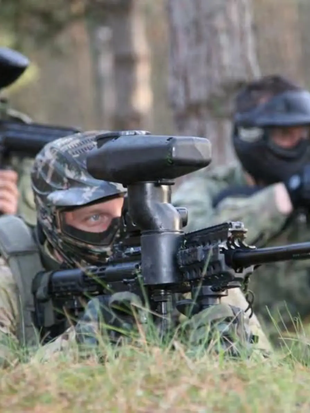 Uczestnik gry terenowej w mundurze i masce celujący z karabinka paintballowego zza trawy