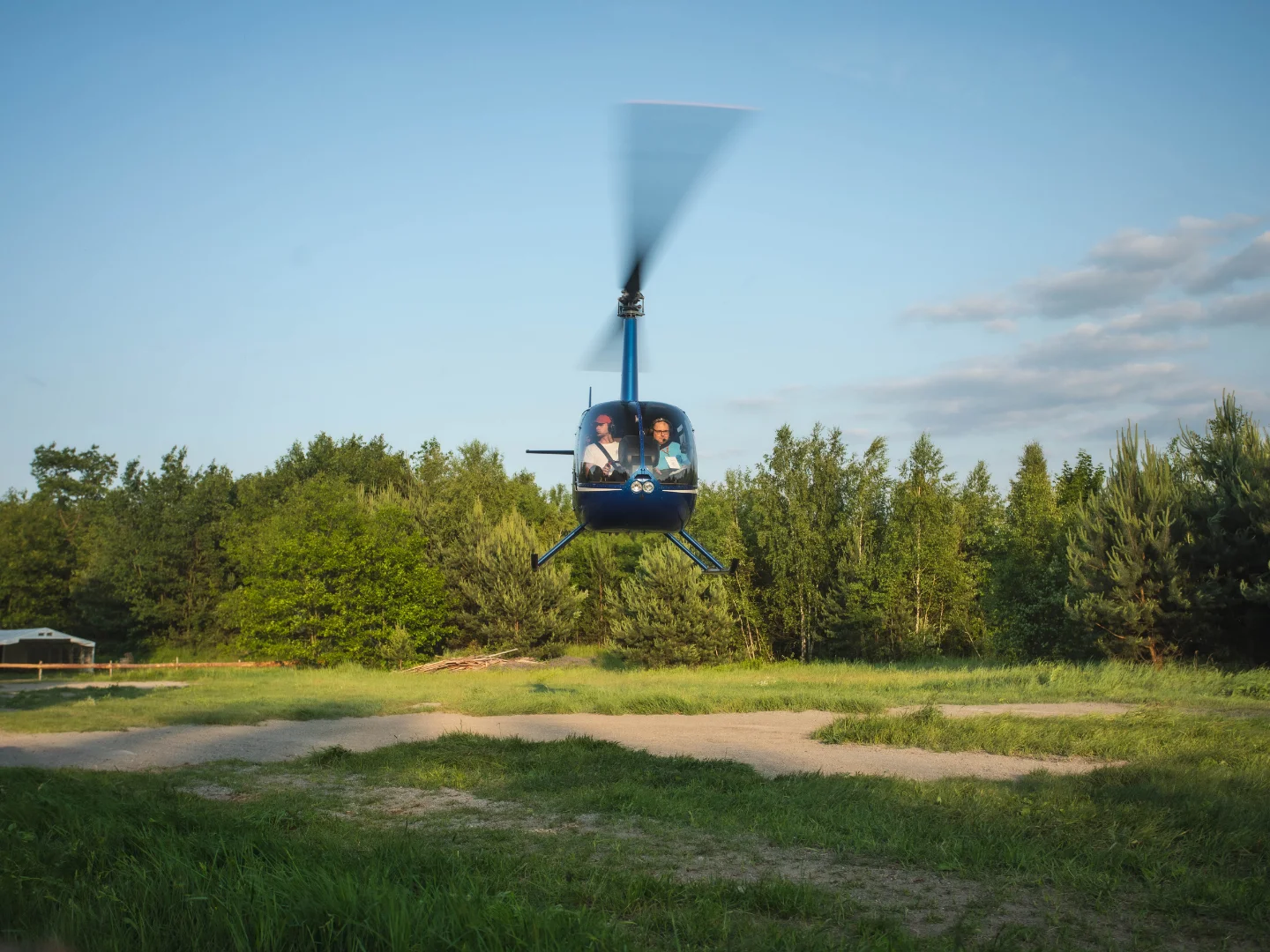 Niebieski helikopter unoszący się nisko nad zieloną polaną na tle drzew