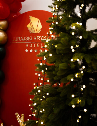 Rozświetlona choinka bożonarodzeniowa na tle ścianki z logo Hotelu Jurajski Kryształ, świąteczne dekoracje wnętrza